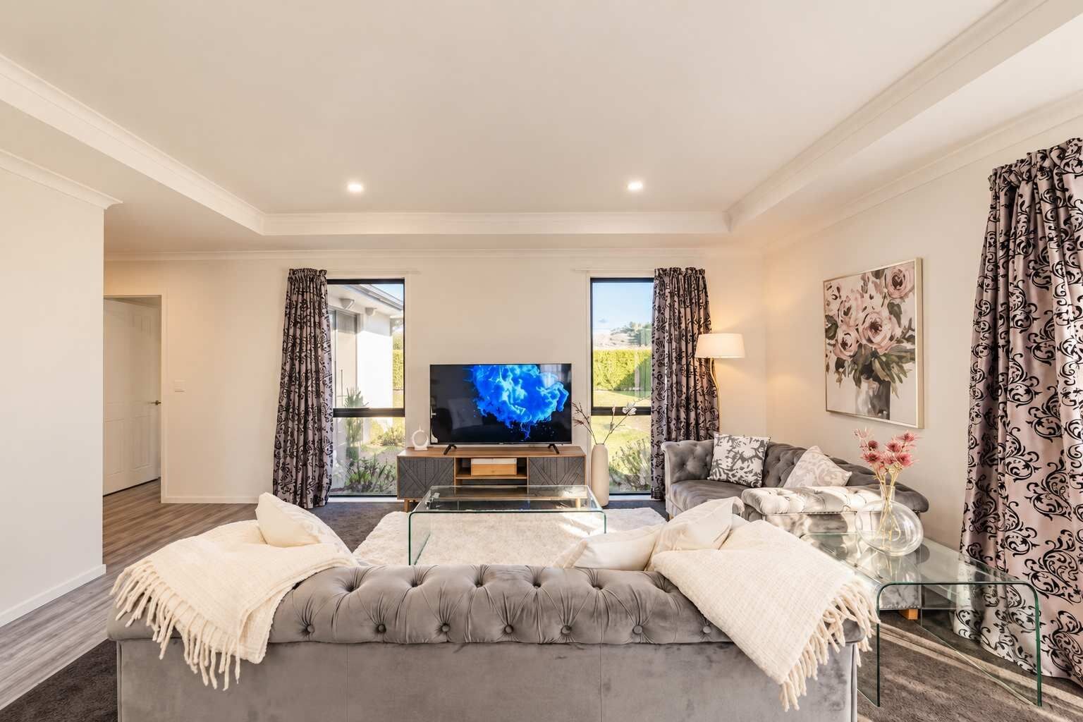 A modern living room with a large flat-screen TV, a grey tufted sofa, a glass coffee table, and a floral painting on the wall.