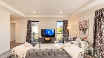 A modern living room with a large flat-screen TV, a grey tufted sofa, a glass coffee table, and a floral painting on the wall.