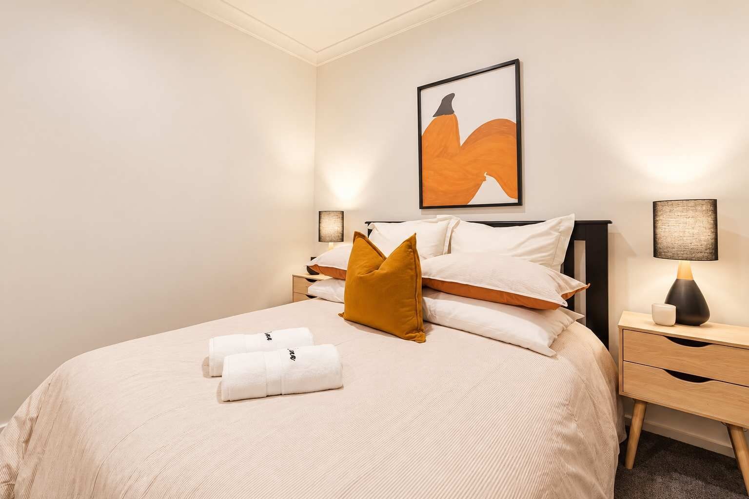Ein ordentlich bezogenes Bett mit Kissen und Handtüchern, ein Nachttisch mit Lampe sowie ein gerahmtes Kunstwerk an der Wand.