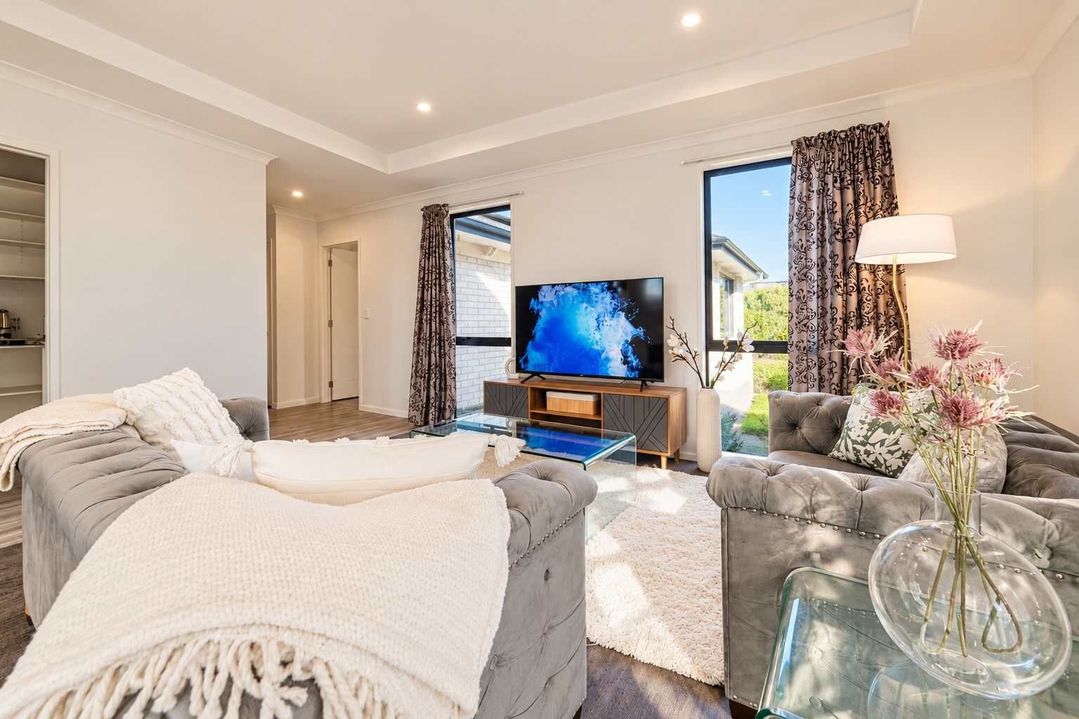 Una sala moderna con un sofá, un sillón y una mesa de centro de vidrio. Un televisor de pantalla plana está montado en la pared y hay una ventana con cortinas.