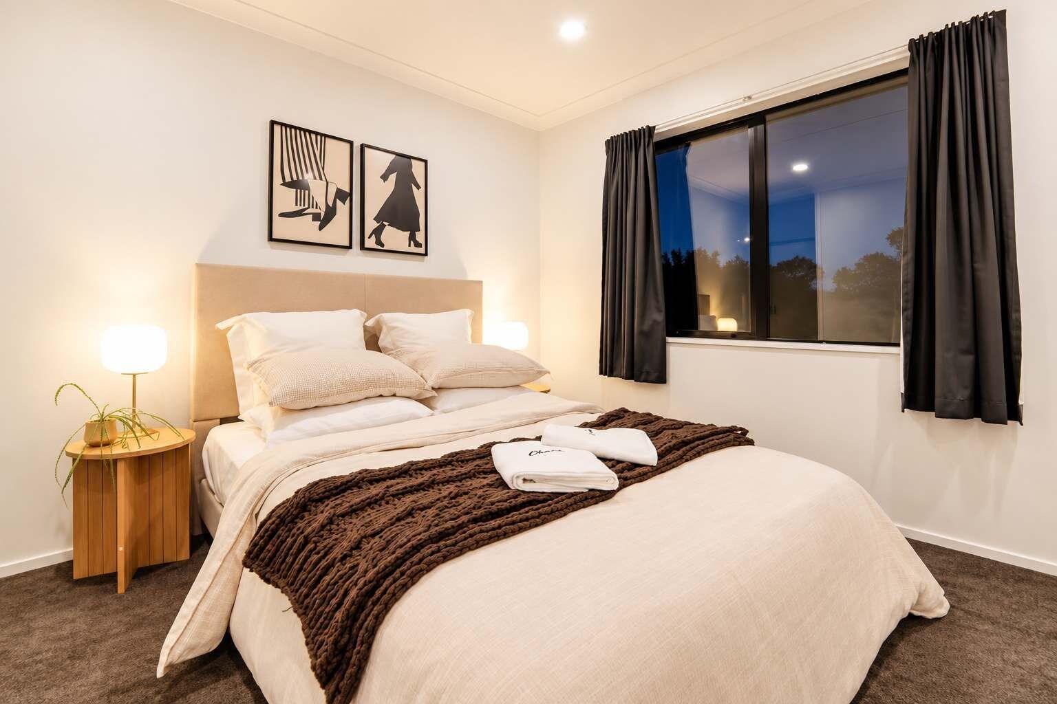 A hotel room with a bed, bedside lamps, a window with curtains, and artwork on the wall.