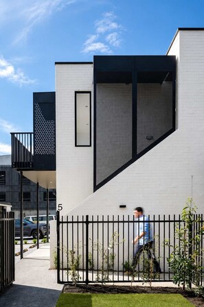 A modern building with a white facade, a black vertical element, and a black fence with greenery.