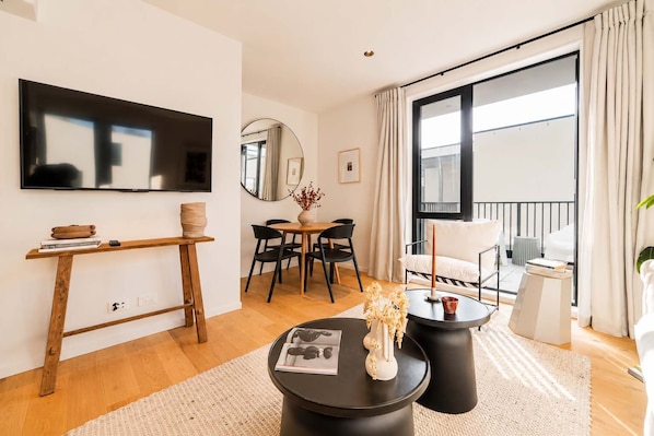 A modern living room with a flat-screen TV, a dining table with chairs, a round coffee table with a magazine, and a balcony with a white chair.