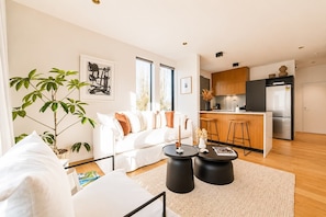A modern living room with a white sofa, a coffee table, and a kitchen area with a refrigerator and bar stools. - Cambridge Terrace | 2BR | Parking + Balcony | CBD (Christchurch)