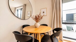 Une table ronde en bois, sur laquelle est posé un vase de fleurs séchées, entourée de chaises noires, dans une pièce dotée d’un grand miroir et de tableaux encadrés accrochés au mur.