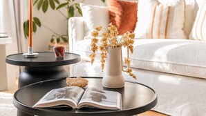 Una sala moderna con un sofá blanco, una mesa de centro con un libro y un objeto decorativo, y un jarrón con flores secas.