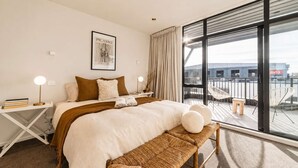 Une chambre d’hôtel moderne avec un grand lit, une table de chevet avec une lampe, une œuvre d’art encadrée au mur et un balcon avec des chaises.