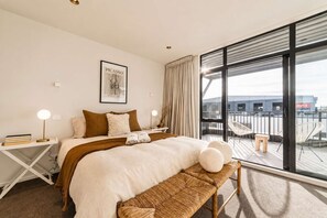 A modern hotel room with a large bed, a bedside table with a lamp, a framed artwork on the wall, and a balcony with chairs.