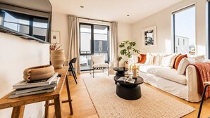 Una sala moderna con sofá, mesa de centro y mesa de comedor con sillas. Hay un ventanal con cortinas, una planta y vistas al exterior.