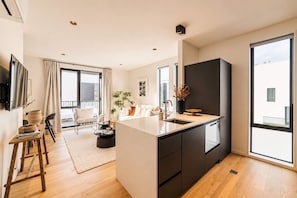 A modern kitchen with a central island, a dining area with a wooden table and chairs, and a living area with a sofa and a coffee table. - Cambridge Terrace | 2BR | Parking + Balcony | CBD (Christchurch)