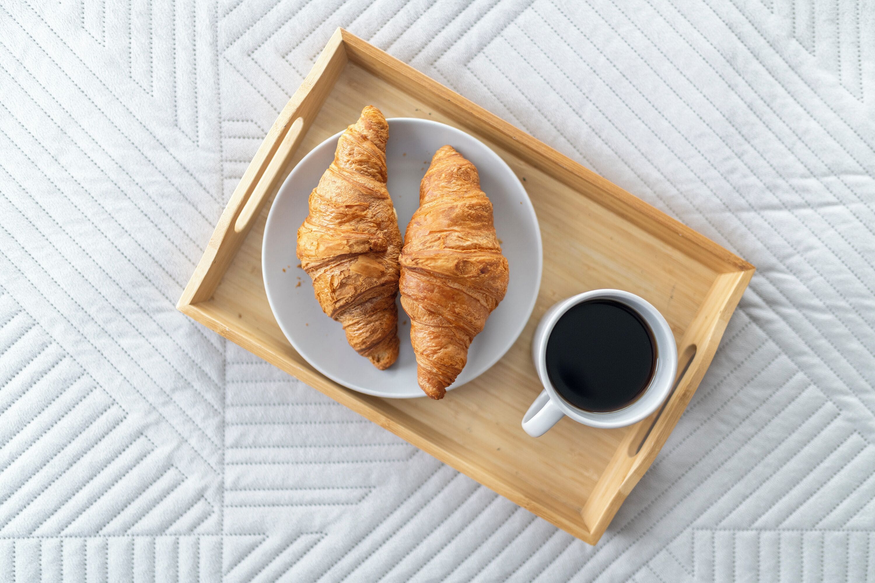 A wooden tray with two croissants and a cup of coffee on a white quilted surface.