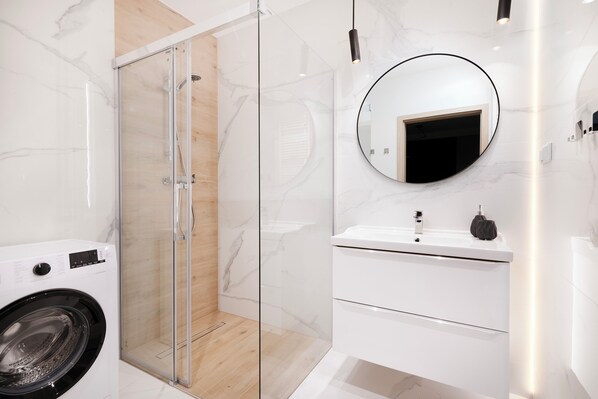 A modern bathroom with a glass shower enclosure, a built-in washing machine, and a white vanity with a mirror. - EASY RENT Apartments- Kompozytorów 7 (Lublin)