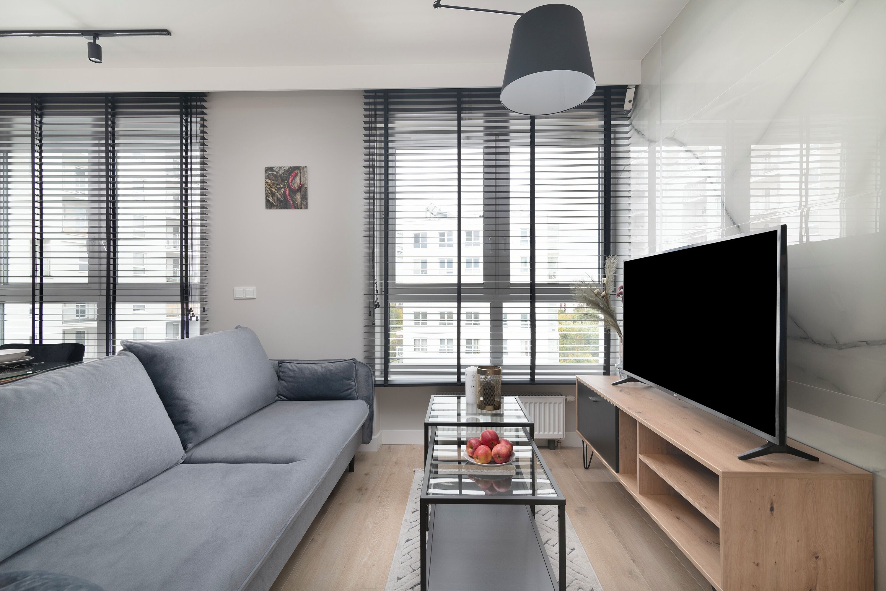 Una sala de estar moderna con un sofá gris, una mesa de centro de vidrio con frutas, un televisor sobre un soporte de madera y ventanas grandes con persianas.