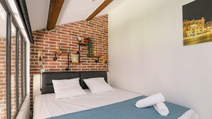 A hotel room with a brick wall, a bed with white linens, a blue bedspread, and a painting of a cityscape.