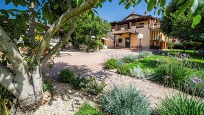 Una casa de dos pisos con un balcón, rodeada por un jardín con diversas plantas y un árbol.