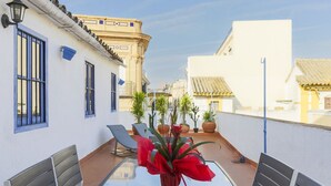 Una terraza en la azotea con una mesa de vidrio, dos sillas, un banco y plantas en macetas.