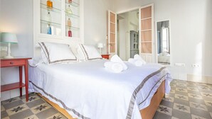 Cama bem arrumada com lençóis brancos e um detalhe azul, um armário de vidro com artigos de higiene, uma mesa de cabeceira vermelha e um piso estampado.