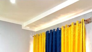 A room with a grey sofa, yellow and blue curtains, and a potted plant. - Silver Stone Cribs (Kampala)