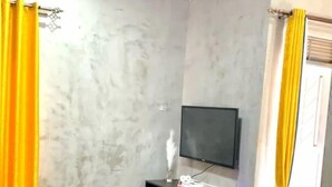 A modern living room with a grey sofa, a flat-screen TV on a black stand, a potted plant, and yellow curtains. - Silver Stone Cribs (Kampala)