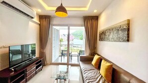 A modern living room with a flat-screen TV, a sofa with cushions, a glass coffee table, and a large window with curtains. - 6-49 - 1 Bdr Condo with Balcony and shared Pool (Tambon Patong)
