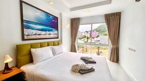 A hotel room with a bed, a painting of the ocean, a window with curtains, and a lamp on the nightstand. - 6-49 - 1 Bdr Condo with Balcony and shared Pool (Tambon Patong)