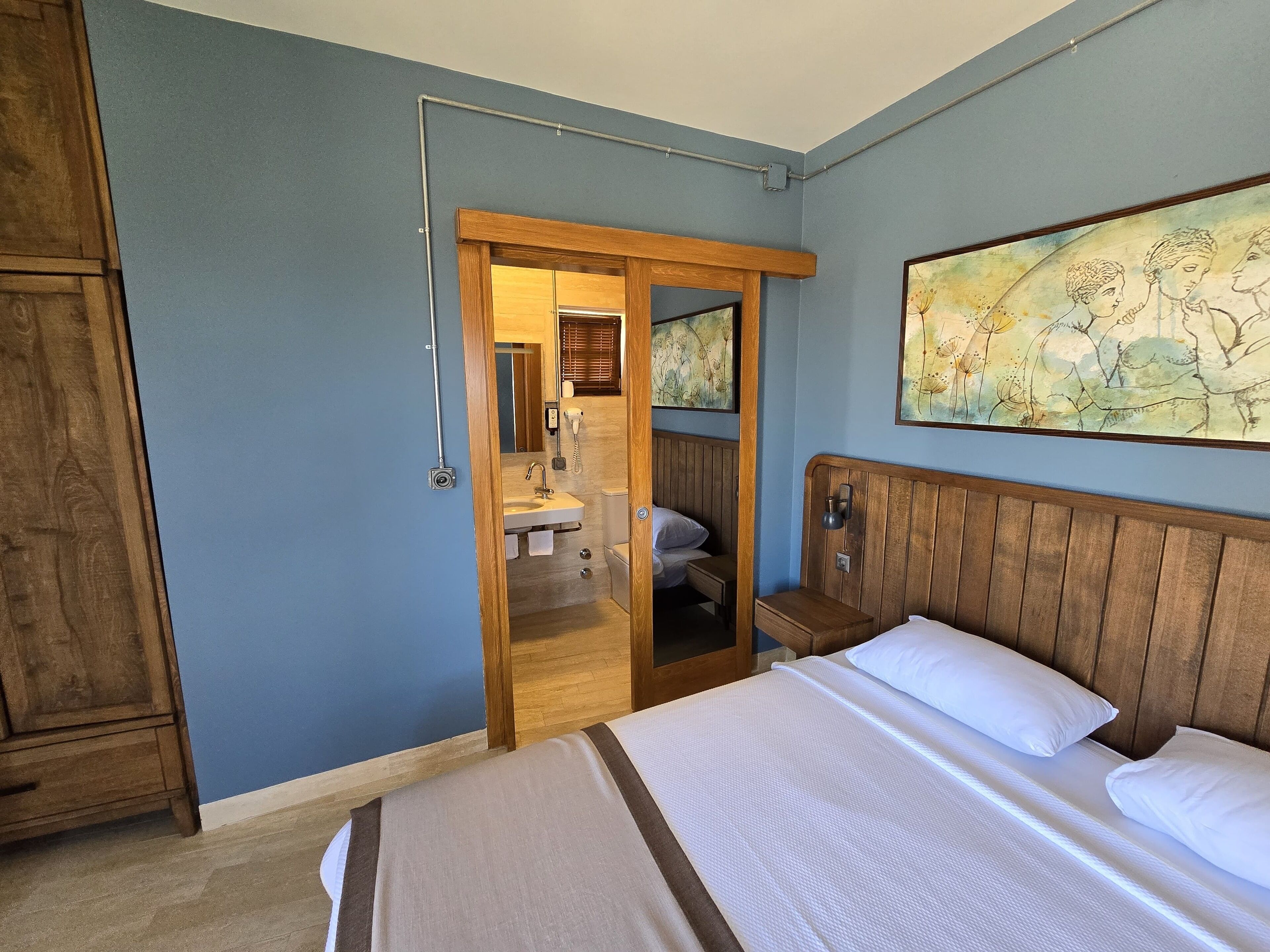 A bedroom with a wooden headboard, a large bed with white bedding, a wooden wardrobe, and a bathroom visible through an open door.