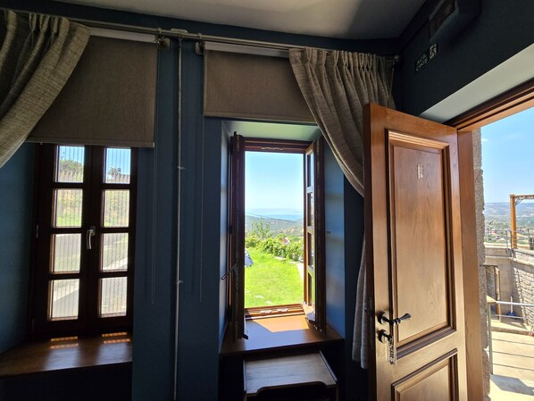 A room with a large window, a wooden door, and a view of a green landscape. - Hane Assos Hotel (Ayvacik)