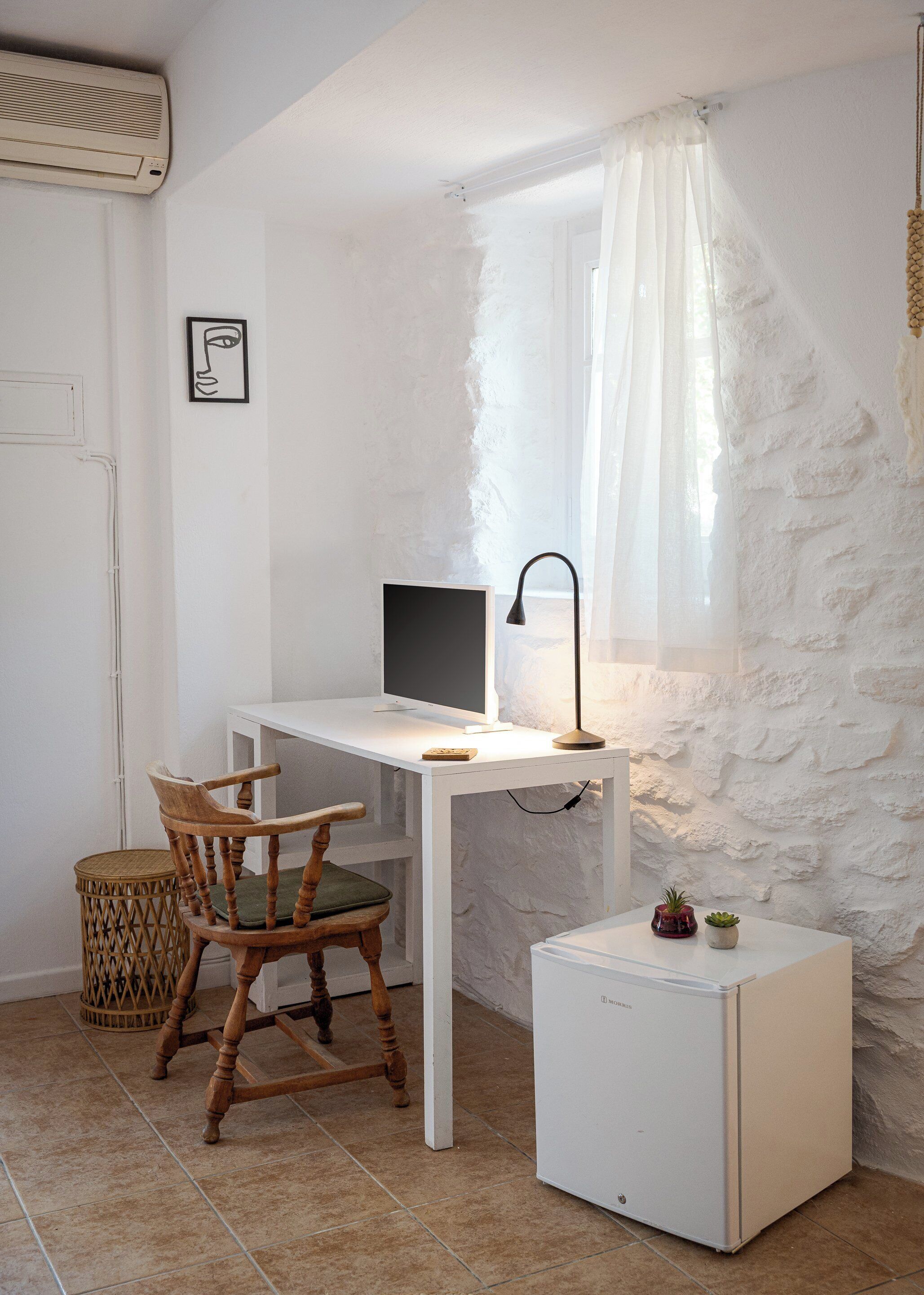 Habitación compacta con escritorio blanco, computadora, silla, refrigerador pequeño y ventana con cortinas de gasa.
