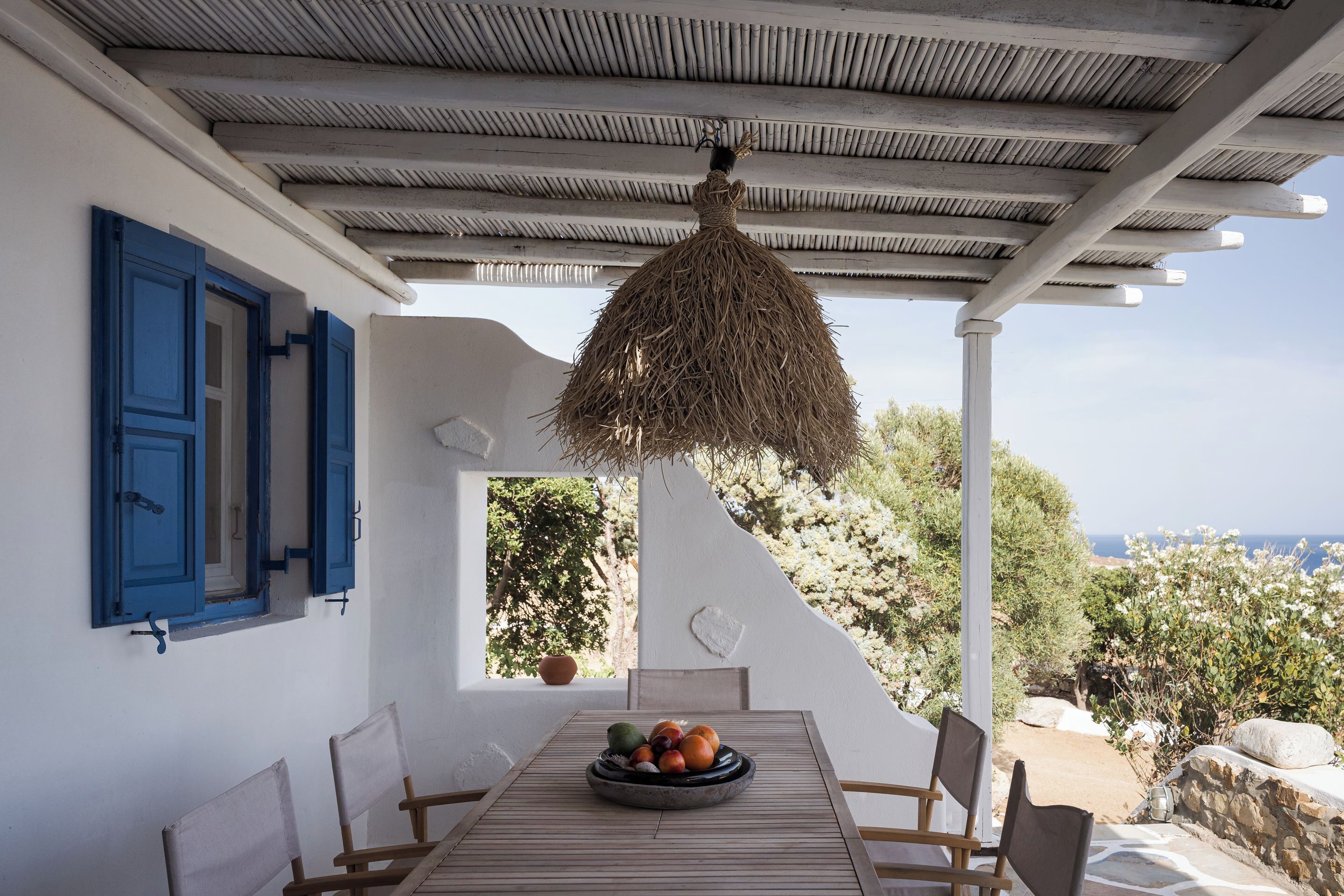 Un tavolo con una ciotola di frutta, pareti bianche, persiane blu e vista sul mare.
