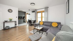 A modern living room with a grey sofa, a small coffee table, and a grey ottoman. There is a dining area with a table and chairs, and a kitchen in the background. - Modern Apartment | Private Parking and Balcony (Konstancin-Jeziorna)