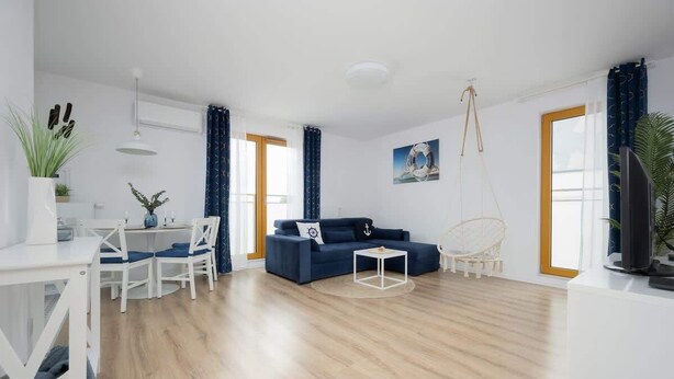 A modern living room with a blue sofa, white dining table, and chairs. There is a TV on a white stand, a hanging plant, and a painting on the wall. - Charming Apartment near Warsaw | Parking, Air Conditioning (Konstancin-Jeziorna)