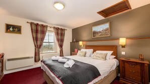 A neatly made bed with a dark blanket and white linens, a wooden nightstand with a lamp, and a window with plaid curtains. - The Red Admiral Hotel (Seascale)