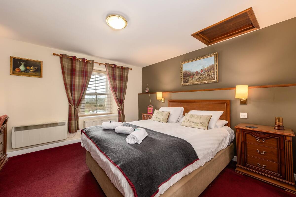 A neatly made bed with a dark blanket and white linens, a wooden nightstand with a lamp, and a window with plaid curtains.