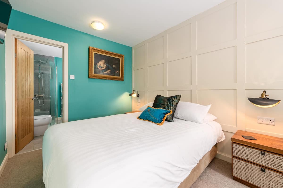 A bedroom with a large bed, a wooden door leading to a bathroom, a framed picture on the wall, and a bedside table with a lamp.