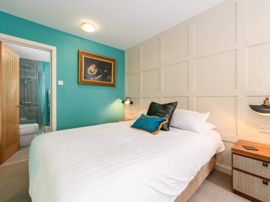 A bedroom with a large bed, a wooden door leading to a bathroom, a framed picture on the wall, and a bedside table with a lamp.