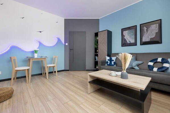 A modern living room with a wooden coffee table, two wooden chairs, a grey sofa with nautical-themed cushions, and wall art depicting maps.