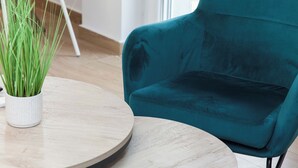 Un fauteuil en velours turquoise est placé à côté d’une table en bois sur laquelle se trouve un pot de fleurs.