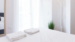 Une chambre d’hôtel lumineuse et blanche, avec un lit, un miroir, un porte-manteau et une fenêtre ornée de rideaux légers.