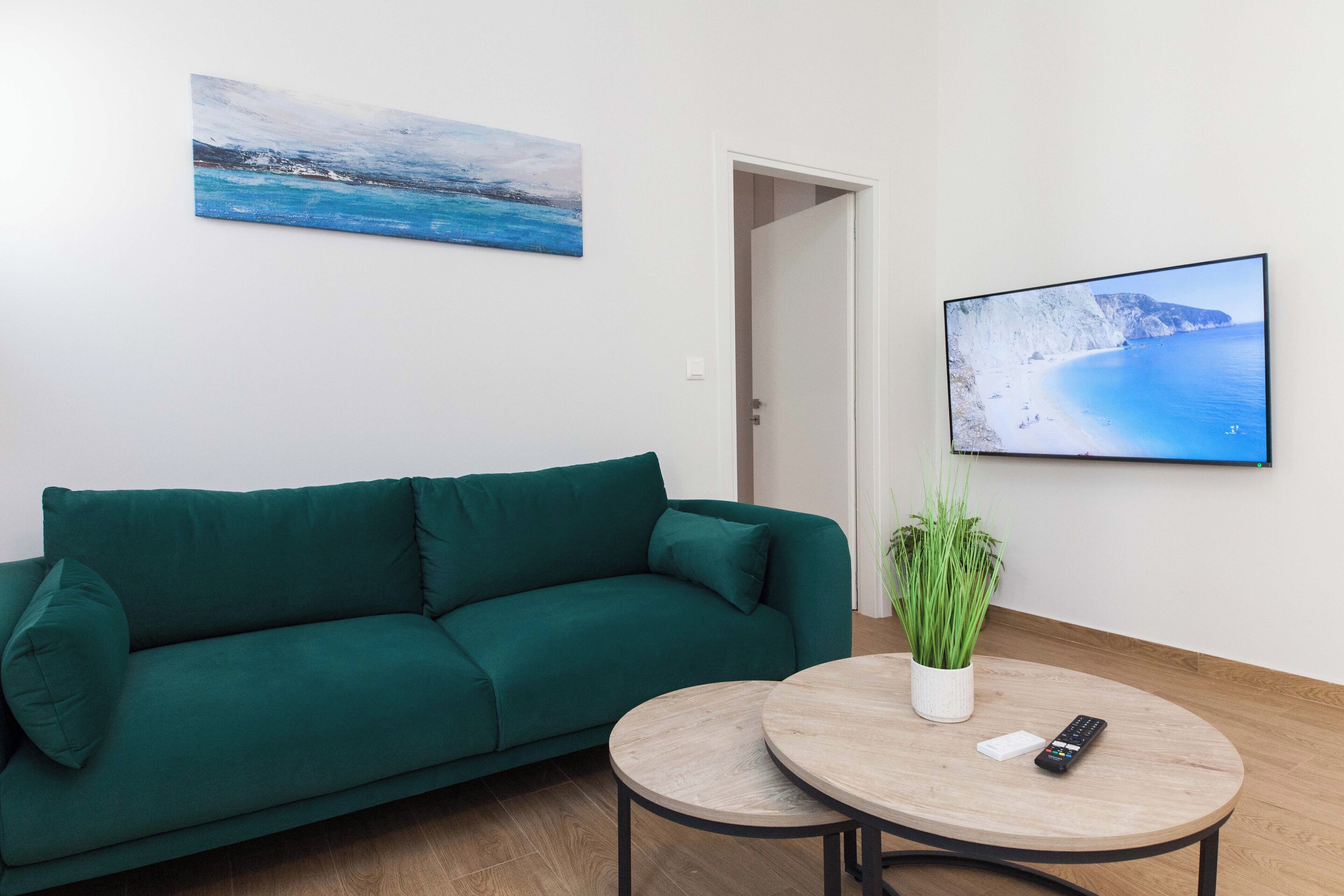 A room with a green sofa, a small round table with a plant, and a TV displaying a coastal scene.