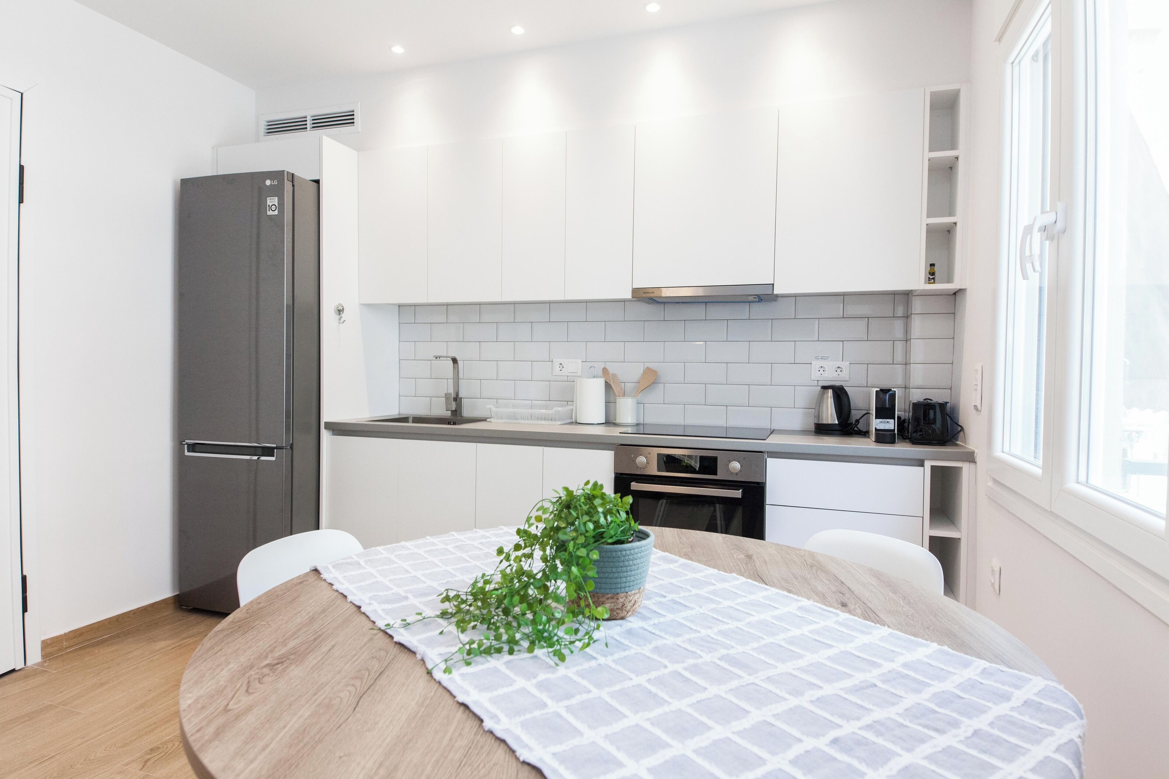 A modern kitchen with white cabinets, a stainless steel refrigerator, a wooden dining table with a checkered tablecloth, and a potted plant.