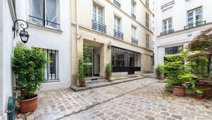 Une cour pavée de pierres, agrémentée de plantes en pot, avec une entrée d’immeuble et un auvent noir.