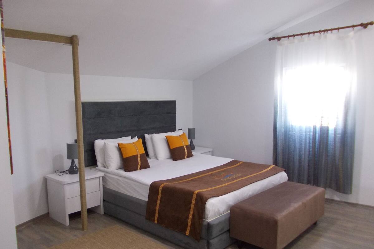 Un dormitorio con cama, mesitas de noche, una banca y una ventana con cortinas.