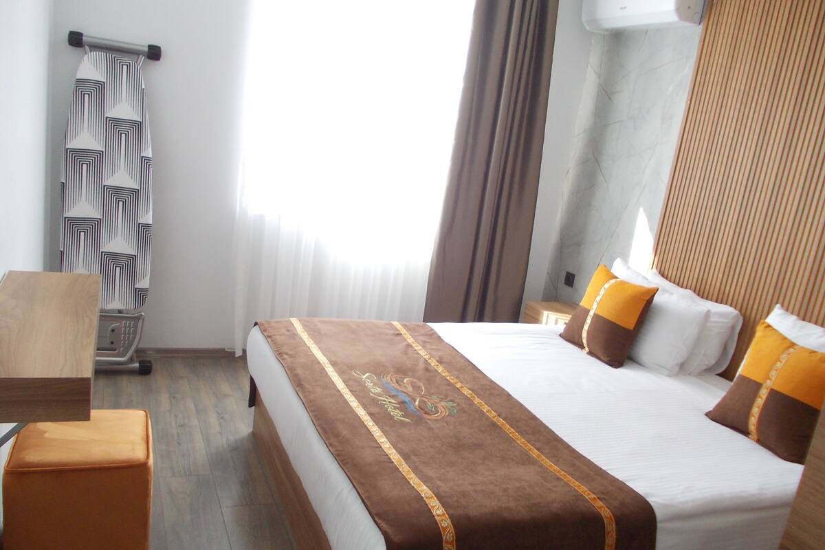 Habitación de hotel con cama, mesita de noche de madera, una pequeña mesa y una ventana con cortinas.