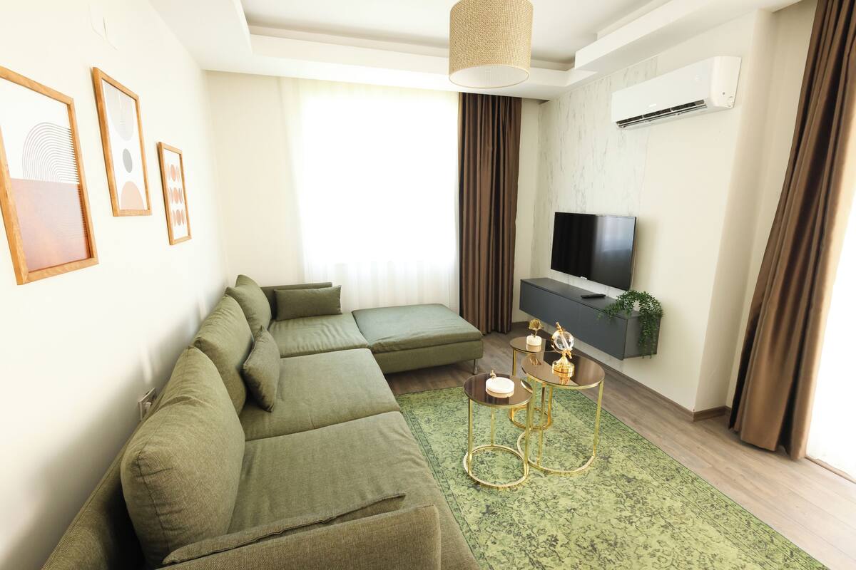 Una sala moderna con un sofá seccional, un televisor de pantalla plana, una mesita auxiliar y una alfombra verde.