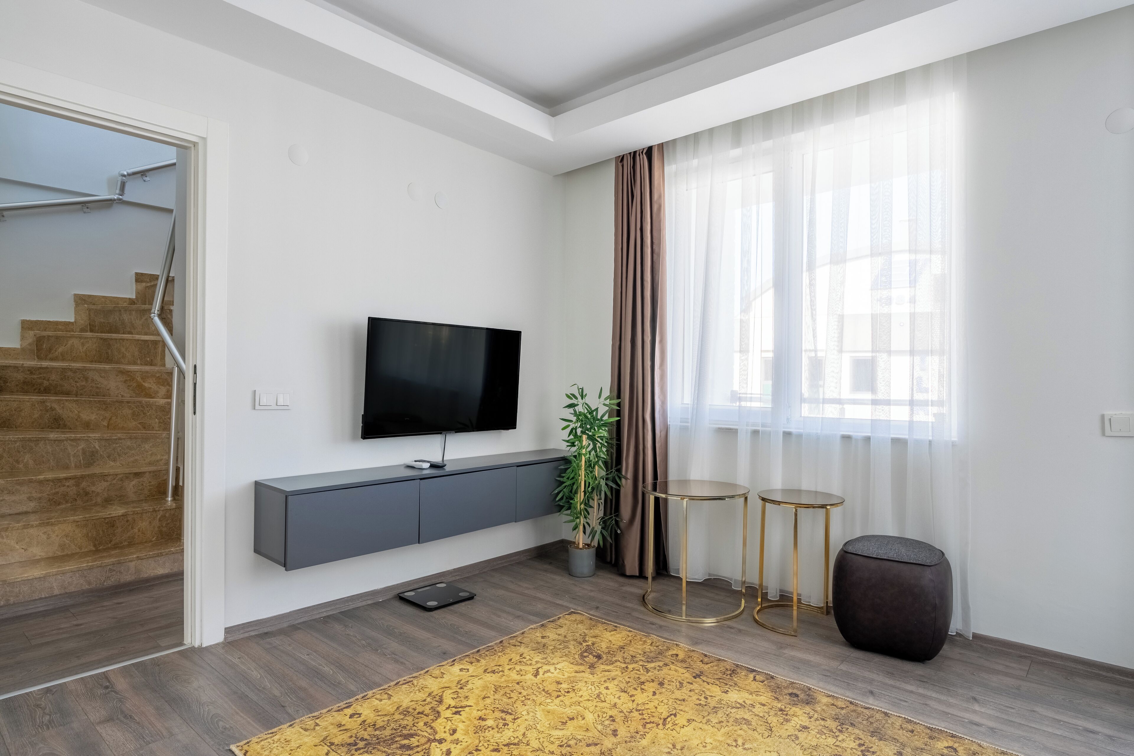 Ein modernes Wohnzimmer mit einem Flachbildfernseher, einer grauen Konsole, einer Topfpflanze, zwei runden Beistelltischen und einer Treppe.