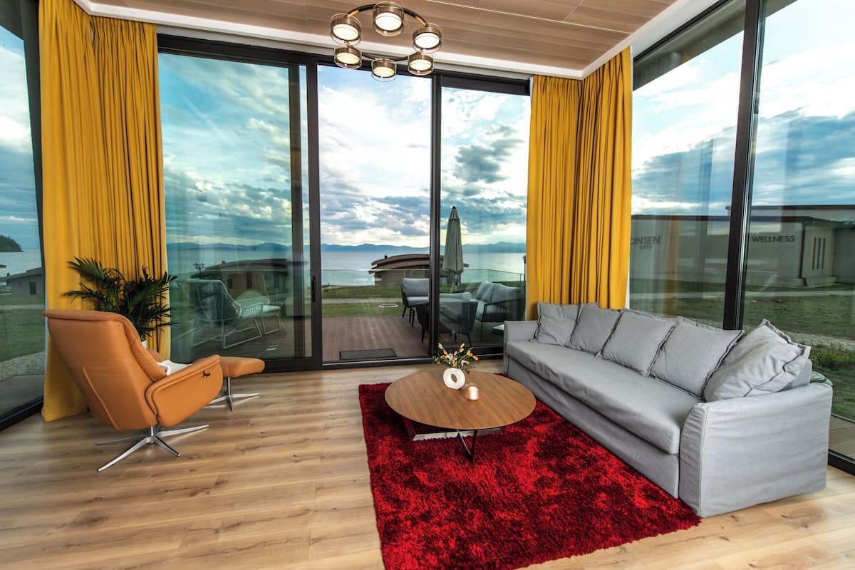 Een moderne woonkamer met een grijze bank, een oranje fauteuil en een rood tapijt. Een rond houten bijzettafeltje met een vaas en kaars staat in het midden. Grote ramen bieden uitzicht op een waterpartij en een ver landschap.
