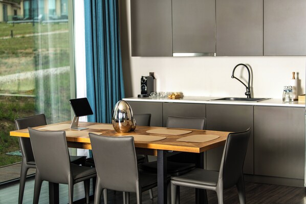 A modern kitchen with a wooden dining table, black chairs, a stainless steel sink, and a view of a green landscape through large windows.