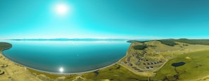 An aerial view of a coastal area with a large body of water, a small settlement with houses and a road, and distant mountains.
