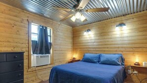 Une chambre à coucher dans une cabane en bois, avec un lit, une commode, un ventilateur de plafond et une fenêtre avec des rideaux.