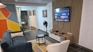 Una sala de estar moderna con sofá, sillón y mesa de centro, un televisor de pantalla plana montado en una pared de madera, y una puerta corrediza de vidrio que da a otra habitación.
